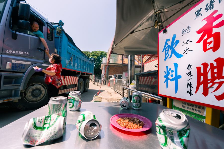 路邊香腸攤前，卡車司機探出車窗與紅衣攤主交談，桌上散落啤酒罐與一盤滷味