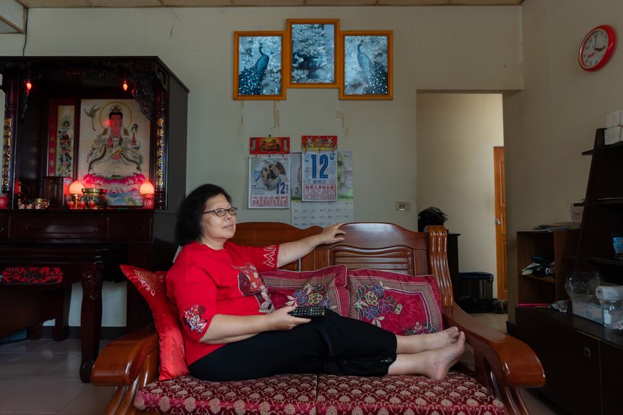 神明廳前，紅衣女子側坐在木沙發上握著遙控器看電視