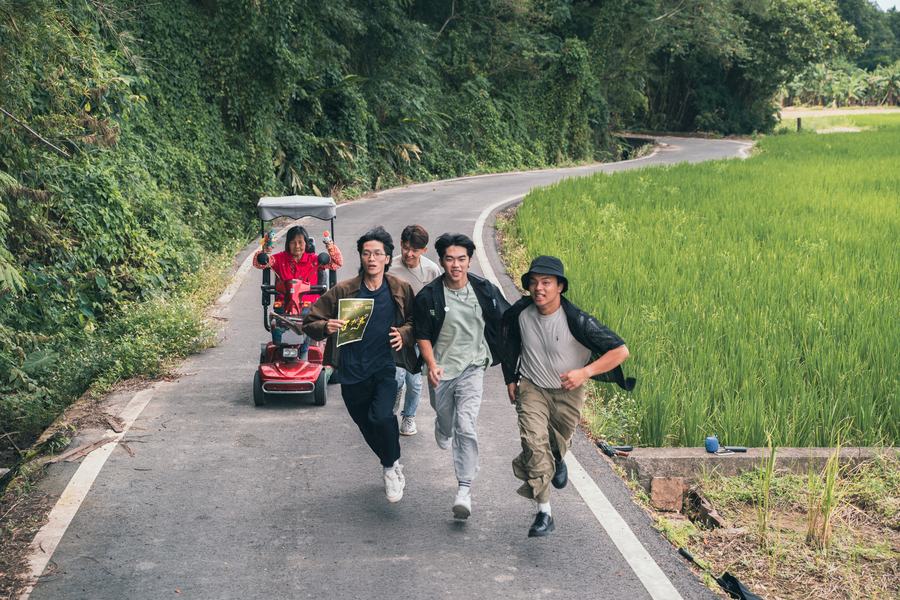 鄉間產業道路上四名年輕人奔跑，後方阿婆騎電動代步車拿著水槍追趕