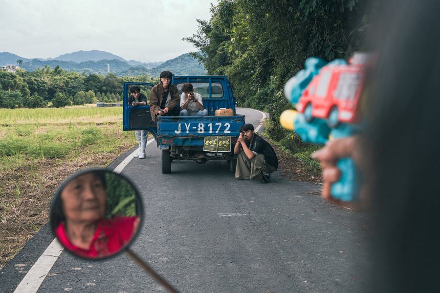 藍色小貨車旁年輕人持水槍對峙，前景後照鏡映出紅衣阿婆的臉