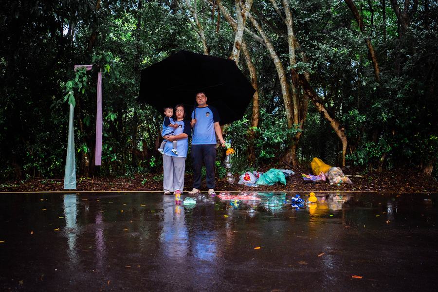 雨後林道旁一家三口撐著黑傘合影，地上散落泡泡與玩具
