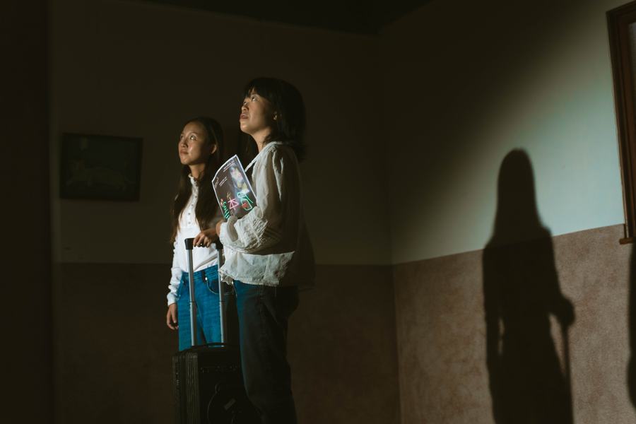 昏暗房間裡兩名女子拉著行李箱抬頭仰望，牆上映出長長的影子