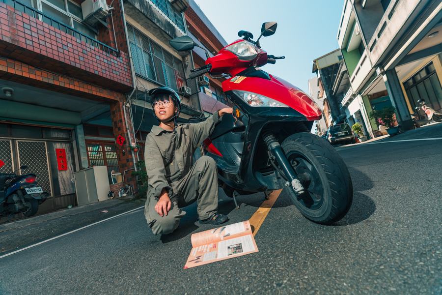 街邊紅色機車旁，穿著工作服與安全帽的女子蹲下看著地上的刊物