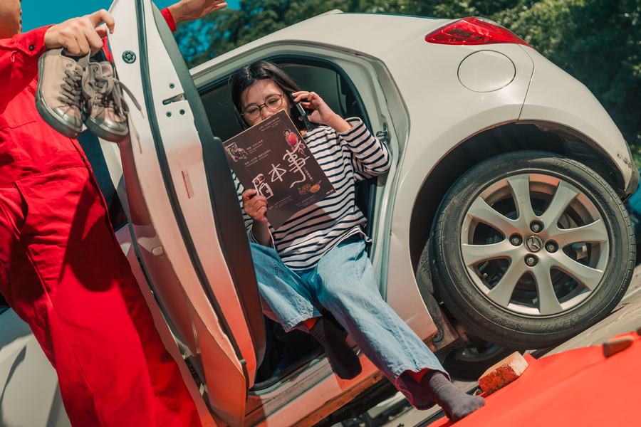 拖吊車上，女子坐在白色轎車駕駛座邊講電話手拿刊物，紅衣男子在旁提著球鞋