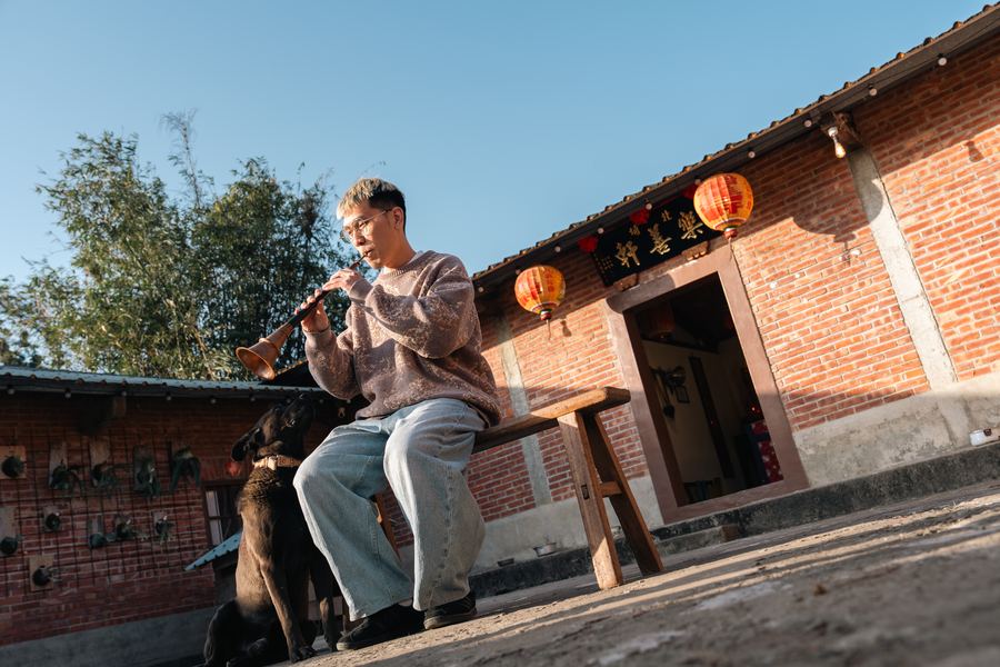 男子在紅磚三合院前吹奏樂器，黑狗坐在腳邊