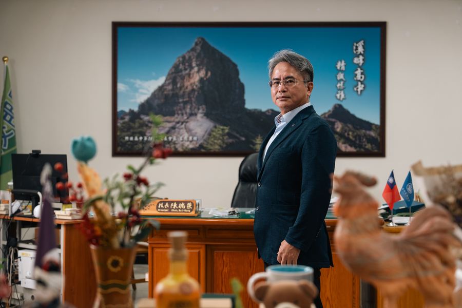 校長站在辦公室內，身後掛著山景大圖