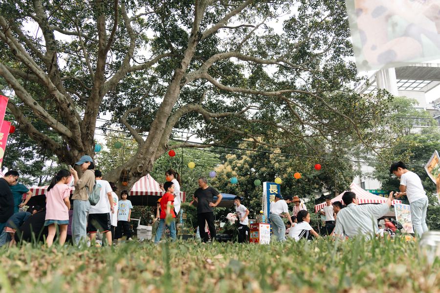 老樹下的村落市集，掛著彩色燈籠，大人小孩在攤位間走動