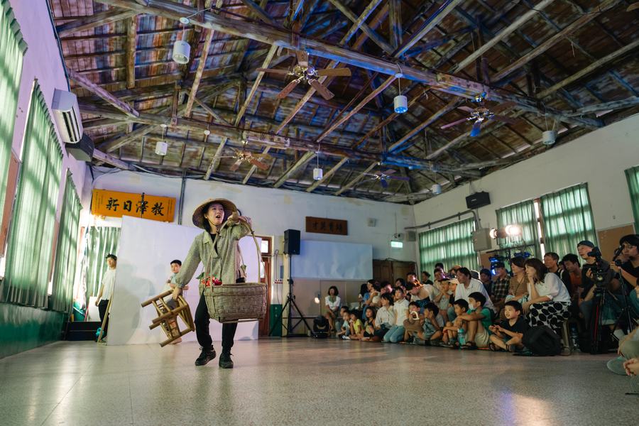 老禮堂裡的戲劇演出，戴斗笠的演員提著竹籃，觀眾席地而坐